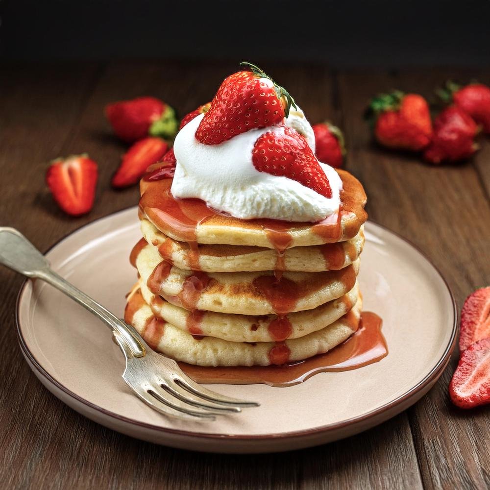 A stack of fluffy sweet cream pancakes topped with whipped cream, fresh berries, and syrup