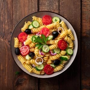 A flat-lay image of fresh pasta salad ingredients, including rotini, tomatoes, cucumbers, olives, and herbs